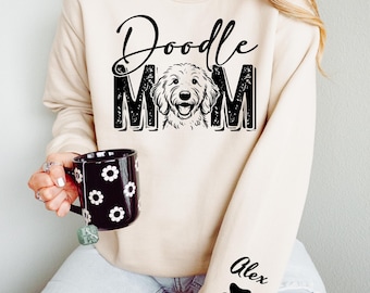 May include: A beige crewneck sweatshirt featuring the black text Doodle MOM with a dog illustration replacing the letter O. The sleeve displays the name Alex above a black paw print. A black mug with white flower patterns is also visible.