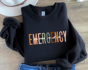 May include: A black long-sleeve sweatshirt featuring the word EMERGENCY in large, multi-colored block letters in shades of orange, tan, and sage green, with the word nurse written in white cursive script layered over the center.