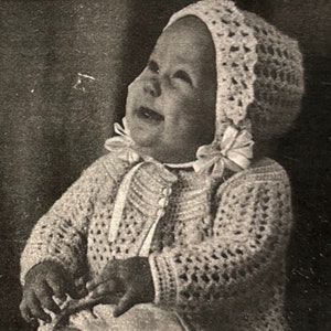 Puede incluir: Una fotografía en blanco y negro de un bebé que lleva un conjunto blanco de ganchillo, que incluye un gorro, una chaqueta y botines. El bebé sonríe y mira hacia arriba.