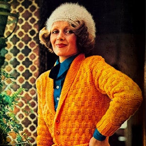 Könnte beinhalten: Eine Frau trägt einen weißen Häkelhut und einen leuchtend orangefarbenen Strickcardigan mit Gürtel und Taschen. Der Cardigan hat einen Schalkragen und einen Knopfverschluss.