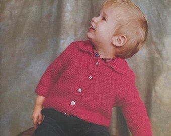 May include: A young child wearing a red knitted cardigan and dark blue shorts sits on a wooden crate. The child is looking up and smiling.