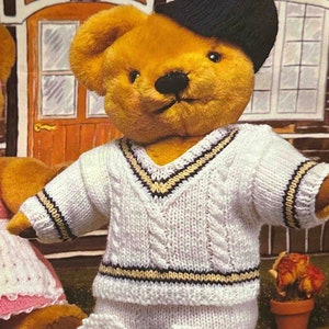 Könnte beinhalten: Ein brauner Teddybär, der einen weißen Strickpullover mit schwarzen und cremefarbenen Streifen, eine weiße Strickhose mit Taschen und eine schwarze Baskenmütze trägt.