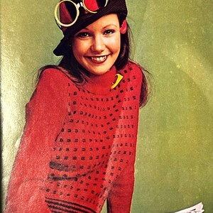 May include: A woman wearing a red sweater with a black and white geometric pattern, a black and red hat with goggles, and a yellow skirt. She is holding a magazine.