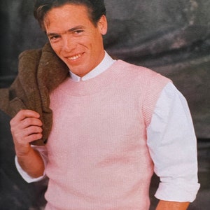 May include: A man wearing a pink knitted vest over a white shirt. He is holding a brown jacket over his shoulder.
