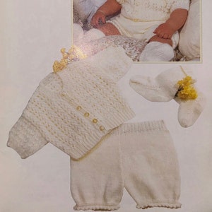 May include: A white knitted baby outfit with a cardigan, pants, and booties. The outfit is made with a delicate stitch pattern and has button closures. The outfit is suitable for babies 18 to 22 inches.