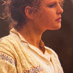 May include: A woman with curly red hair is wearing a cream-colored cable knit sweater with blue and red stripes. The sweater has a v-neck and a button closure.