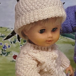 May include: A doll wearing a white knitted hat with a white pom-pom, a white knitted sweater, purple pants, and red shoes. The doll is holding a red bag with a brown buckle.