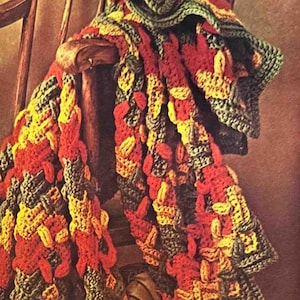 Puede incluir: Una manta de ganchillo con un patrón de hojas en tonos de verde, rojo, amarillo y naranja. La manta está drapeada sobre una silla de madera y hay manzanas y uvas en primer plano.