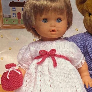 May include: A doll wearing a white knitted dress with pink accents and a pink knitted bag. The doll has brown hair and blue eyes. The doll is wearing pink shoes.