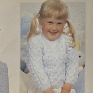 Könnte beinhalten: Ein hellblauer Strickpullover mit Zopfmuster. Der Pullover hat einen runden Ausschnitt und lange Ärmel.