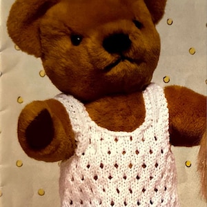 May include: A brown teddy bear wearing a white crocheted tank top and purple and white striped shorts.