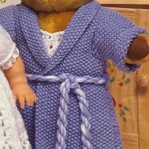 May include: A brown teddy bear wearing a purple knitted robe with a white and purple braided belt.