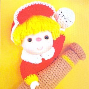 May include: A crochet doll with yellow hair, a red hat, and a red outfit, holding a brown baseball bat and a white ball. The word "Popcorn" is at the top, and the text "Brought to you directly from 'Cupcake Corner'" is at the bottom.