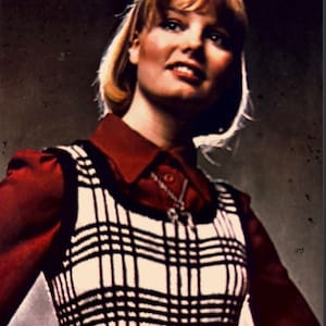 May include: A woman wearing a red collared shirt with a black and white plaid vest. She is wearing a black belt with a gold buckle and red and black plaid pants.