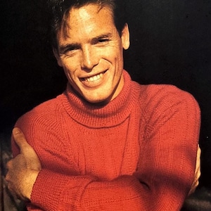 May include: A man wearing a red turtleneck sweater with a ribbed texture. He is smiling and has his arms crossed.