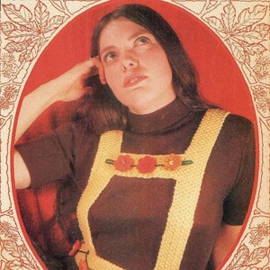 May include: A brown turtleneck sweater with a yellow crocheted vest featuring a floral design with red, orange, and pink flowers. The vest has a criss-cross strap design in the back.