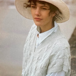 May include: A woman wearing a white knitted sleeveless sweater with a cable knit pattern. She is also wearing a white hat and a white shirt with thin blue stripes.