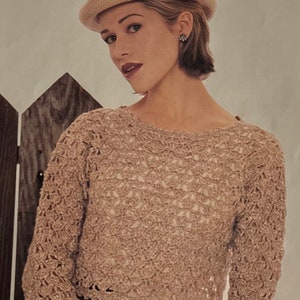May include: A beige crocheted sweater with long sleeves and a round neckline. The sweater is cropped and has a delicate lace trim. The model is wearing a brown hat and a dark skirt.