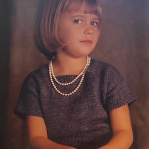 May include: A young girl with short blonde hair wearing a gray sweater and a pearl necklace. She is sitting with her hands folded in her lap.