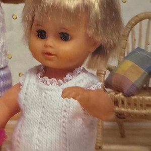 May include: A blonde doll with blue eyes wearing a white crocheted sleeveless top and a pink diaper. The doll has hair rollers in her hair and is sitting on a wooden floor.