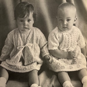 May include: Two babies wearing matching white knitted dresses with a lace trim. The dresses have a ribbon tied at the waist. The babies are sitting on a textured surface.