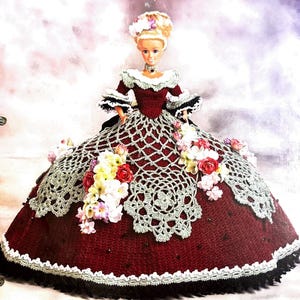 May include: A crocheted doll dress with a burgundy bodice and a white lace overlay. The dress is decorated with white and pink flowers. The doll is wearing a black choker necklace and has blonde hair.