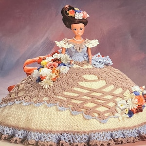 May include: A crocheted doll dress with a brown and white lace pattern. The dress has a blue trim and is decorated with a floral bouquet.