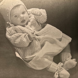 May include: A baby wearing a knitted white hat and matching cardigan with a white dress underneath. The baby is sitting on a brown surface.