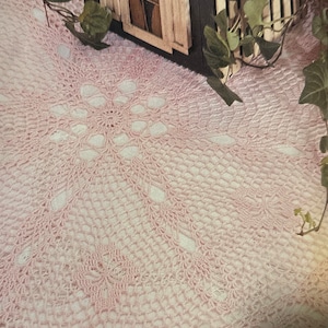 May include: A pink crocheted doily with a delicate floral design. The doily is laid out on a white surface with a small wooden house and green ivy in the background.
