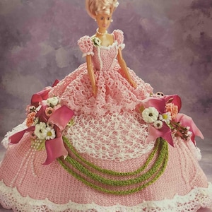 Può includere: Una bambola con un abito rosa all'uncinetto con finiture in pizzo bianco e un'acconciatura abbinata. L'abito presenta un corpetto dettagliato, una gonna ampia ed è adornato con fiocchi rosa, fiori e dettagli di perline verdi. La bambola ha una collana di perle.