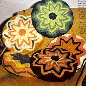 Pode incluir: Quatro almofadas de crochê em forma de flor em várias cores, incluindo laranja, verde, marrom e amarelo. As almofadas estão dispostas em uma superfície marrom.