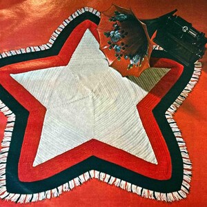May include: A white crocheted star-shaped rug with red and black borders and white fringe. The rug is on a red surface.