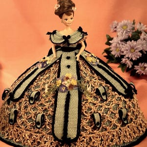 May include: A doll wearing a crocheted dress with a green and brown pattern. The dress has black trim and a black bow at the neckline. The doll has brown hair and blue eyes.