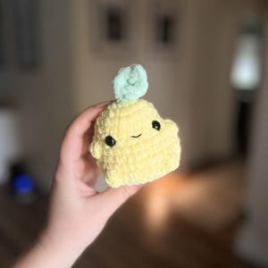 May include: A small crocheted plush toy shaped like a yellow ghost with a green flower on top. The ghost has a smiling face with black eyes.