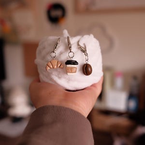 Pode incluir: Três pingentes de comida em miniatura: um croissant, uma xícara de café e um grão de café. Os pingentes são presos a fechos prateados. Os pingentes são segurados contra um fundo branco e fofo.