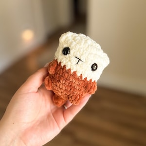 May include: A small, handmade crochet plush toy shaped like a sea otter. The upper portion is cream-colored, while the lower body and small limbs are a rich, textured brown. It features two round, black plastic eyes and a stitched black mouth.