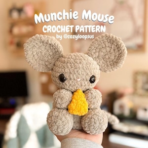 Puede incluir: Un juguete de ratón tejido a ganchillo en tonos grises, sosteniendo una cuña de queso amarillo. El ratón tiene orejas grandes y redondas y ojos negros. El texto en la parte superior dice "Munchie Mouse CROCHET PATTERN by @cozyloopsus."