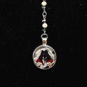 May include: A silver chain necklace with a bat charm, a decorative filigree charm, and a round pendant featuring a hooded figure. The necklace is adorned with green, red, and white beads.