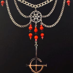 May include: Silver necklace featuring multiple chain strands and red bead details. A decorative silver pendant with a circular design hangs above a copper-coloured cross pendant. The necklace is set against a black backdrop.