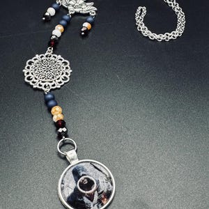 Peut inclure: Un collier en chaîne argentée avec un pendentif rond représentant une figure à capuche. Le collier comporte également un charme en filigrane argenté et un charme avec un motif de papillon. Le collier est orné de perles noires, blanches et rouges.