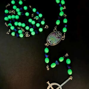Puede incluir: Un rosario con cuentas verdes brillantes, intercaladas con cuentas azul oscuro y negras. El rosario presenta una cruz plateada y un colgante con un diseño de calavera. El fondo es negro.