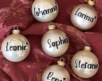 Könnte beinhalten: Sechs weiße Weihnachtskugeln mit goldenen Spitzen und schwarzen Schriftzügen. Die Kugeln sind mit den Namen "Johanna", "Larissa", "Leonie", "Sophia", "Stefanie" und "Rene" verziert.