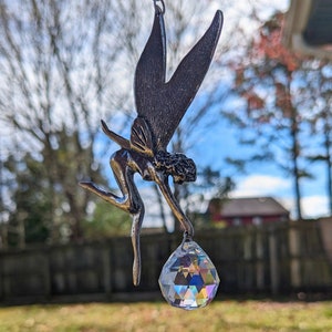 May include: A metal fairy sculpture with outstretched wings and a crystal prism. The fairy is dark gray and appears to be in a graceful pose, reaching down to the faceted crystal. The crystal is multi-colored and teardrop-shaped.