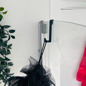 May include: A black shower pouf hanging from a black hook attached to a glass shower door. The hook is attached to a silver shower door track.