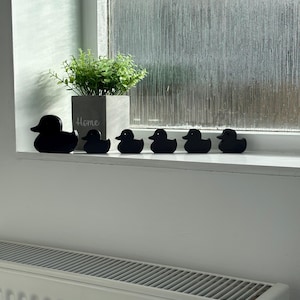 May include: A set of six black duck-shaped figures arranged in a line on a white windowsill. Next to the largest duck is a gray square planter containing a small green plant, with the word Home printed in white script on the front of the planter.