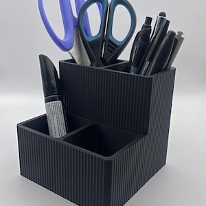 May include: Black desk organiser with four compartments. The organiser has a ribbed texture and holds a pair of scissors, a marker, and several pens.