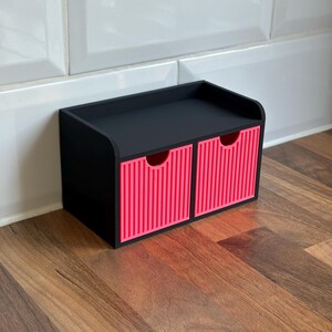 May include: A black desktop organizer featuring two drawers with bright pink, vertically ribbed front panels. The unit has a flat top surface and a rectangular frame, providing a compact storage solution with two circular cutouts for drawer access.