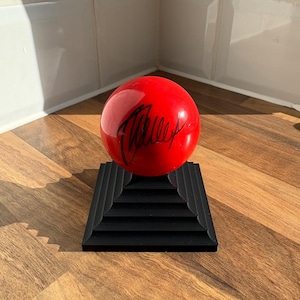 May include: A glossy red ball with a black signature rests on a black, tiered stand. The ball is the focal point, with the signature adding a unique detail. The stand provides a stable base, creating a visually balanced composition. The items are on a wooden surface.