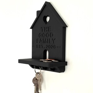 May include: A matte black wall-mounted key holder shaped like a house with a small shelf. The front features a cutout heart, the text "THE GOOD FAMILY" and "EST. 2026" embossed in the center. A set of metal keys hangs from a hook underneath the shelf.
