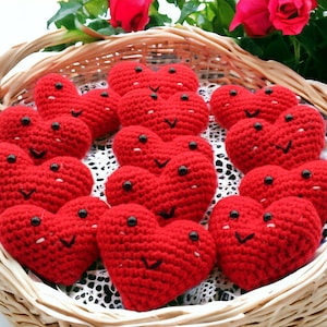 May include: A wicker basket filled with red crocheted hearts, each with black button eyes and a stitched smile. The hearts are arranged on a white lace doily. Red roses are visible in the background.